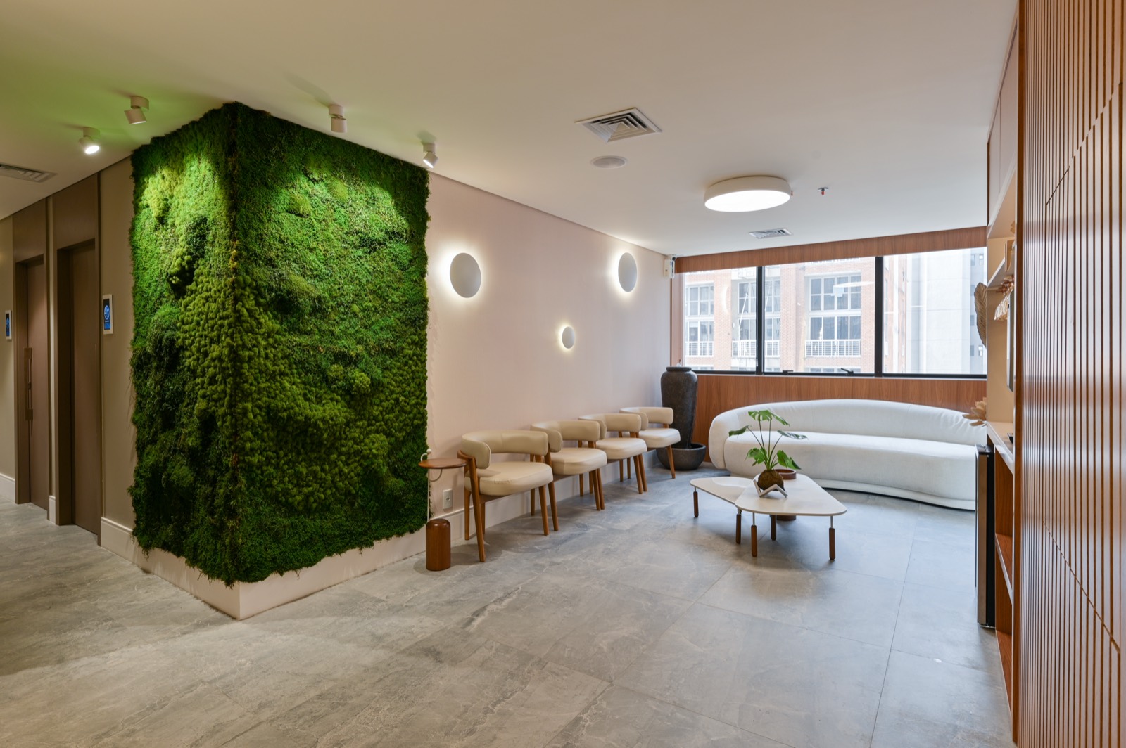 Sala de espera com jardim vertical, poltronas claras e sofá curvo junto à janela